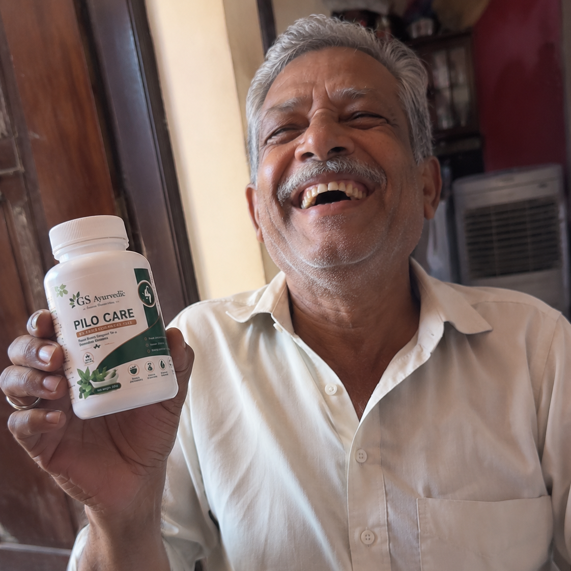 A smiling man holding up a white bottle of GS ayurvedic Pilo Care dietary supplement in front of a home  background.